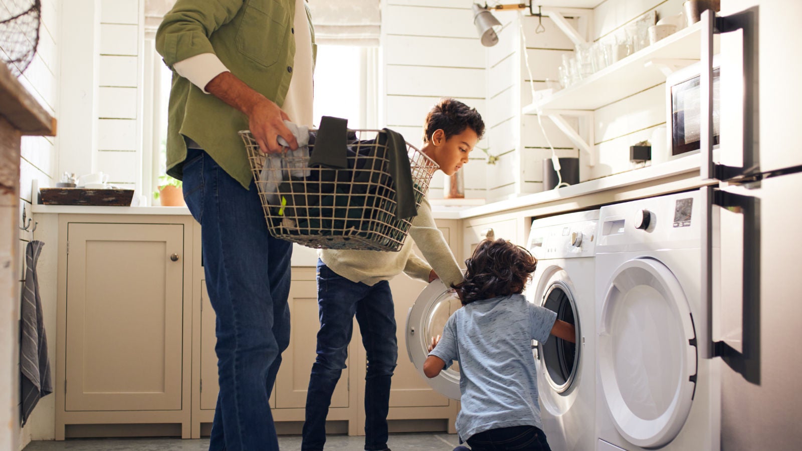 A family doing laundry together in a bright, modern home. A father holds a laundry basket while two children load clothes into a washing machine.