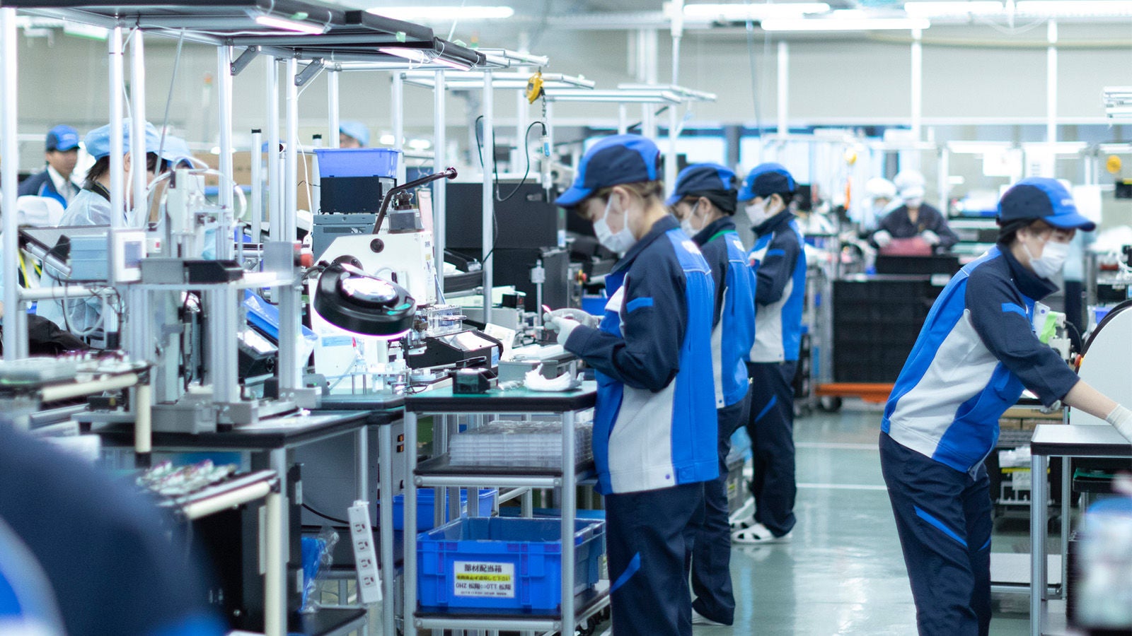 Multiple factory workers in blue uniforms and caps assembling medical devices in a clean manufacturing facility.