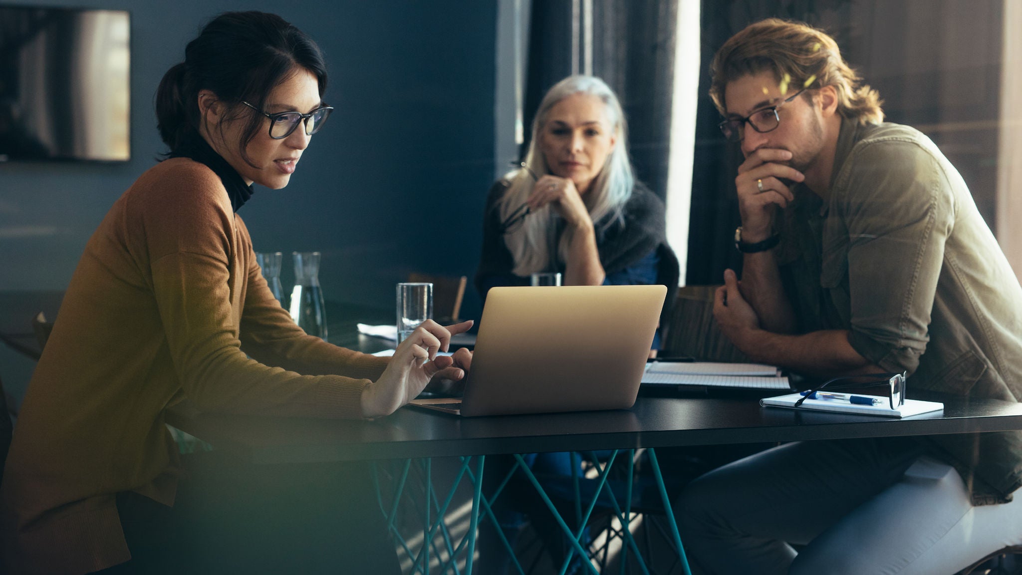 Three colleagues sit around a table in an office, reviewing information on a laptop and discussing work-related matters, with notebooks, pens, and glasses of water nearby.