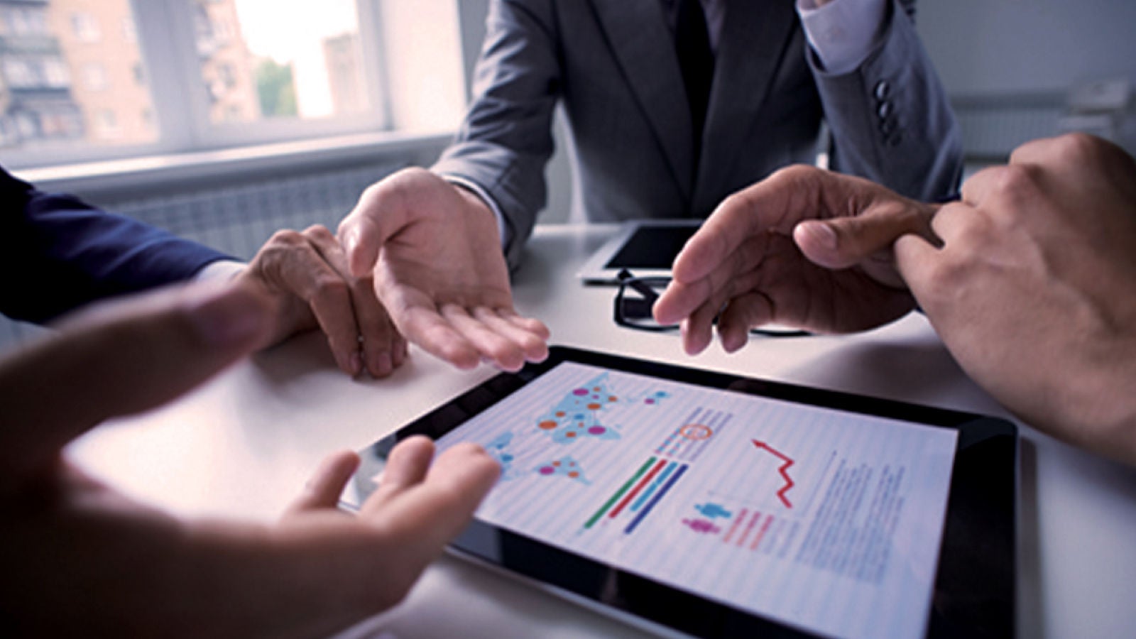 A man seated at a table, using a laptop, gestures with his hand while engaged in conversation.