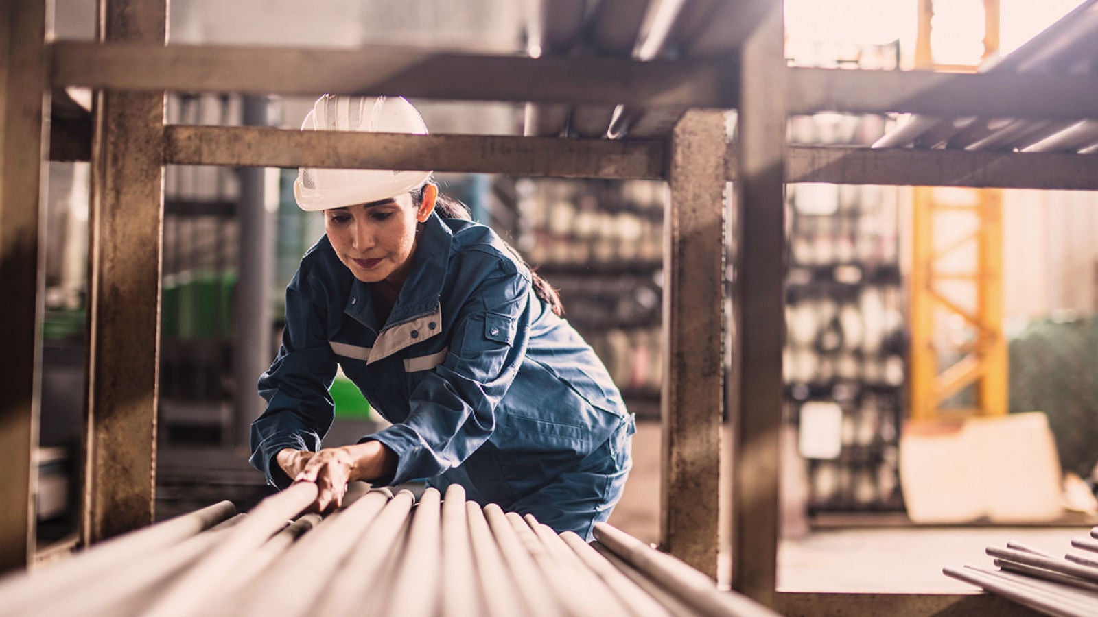 Worker wearing a hard hat and safety uniform arranging metal pipes inside an industrial facility.