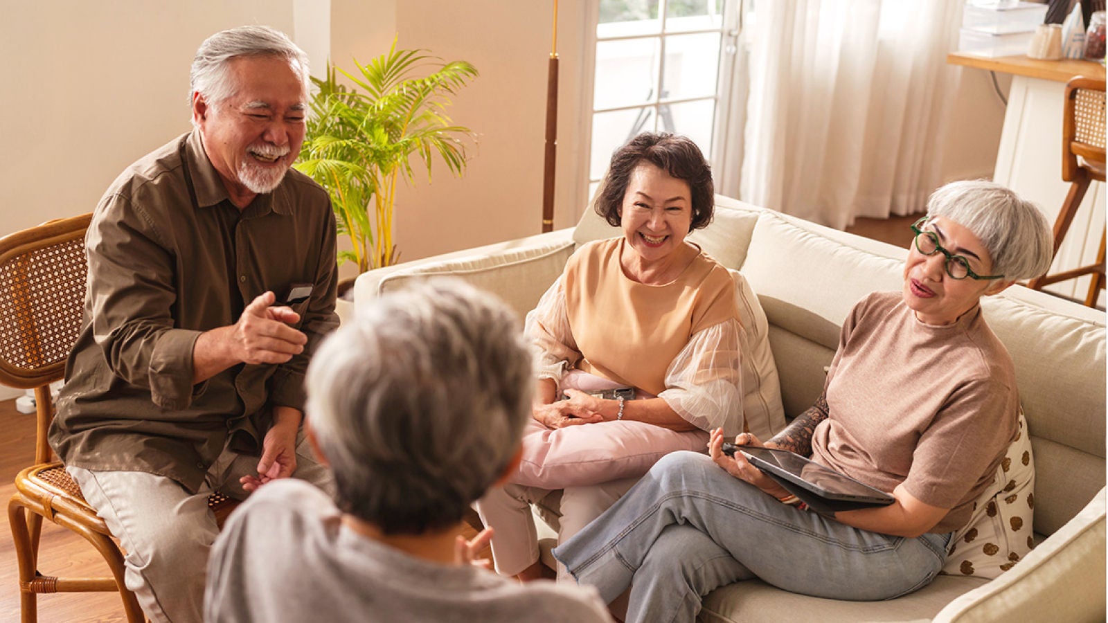Four older adults laughing together in a bright living room, with one person holding a tablet.