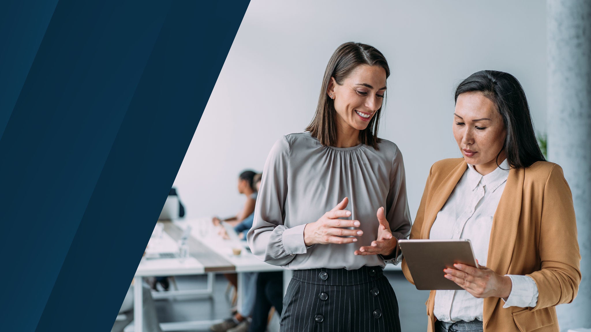 Two business professionals standing in a modern office, smiling and reviewing content on a tablet with colleagues working at desks in the background.