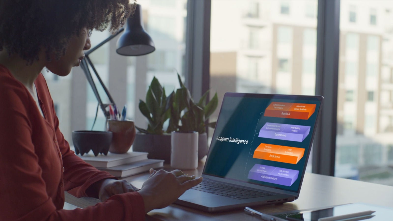 A woman sitting by a window using a laptop displaying an Anaplan Intelligence diagram with AI components.