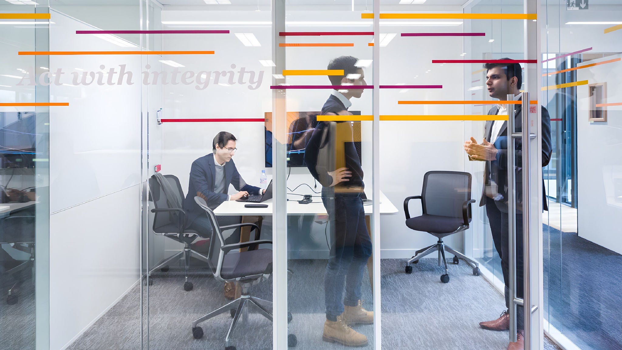 Three men inside a glass-walled office, one seated and working on a laptop while the other two stand and talk. Colored lines and the phrase "Act with integrity" are visible on the glass wall.