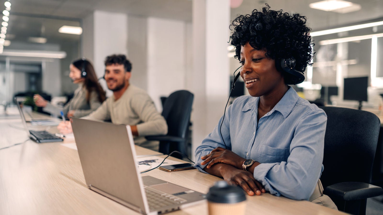 Customer service agent wearing a headset smiling while using a laptop at a desk, with colleagues working in the background.