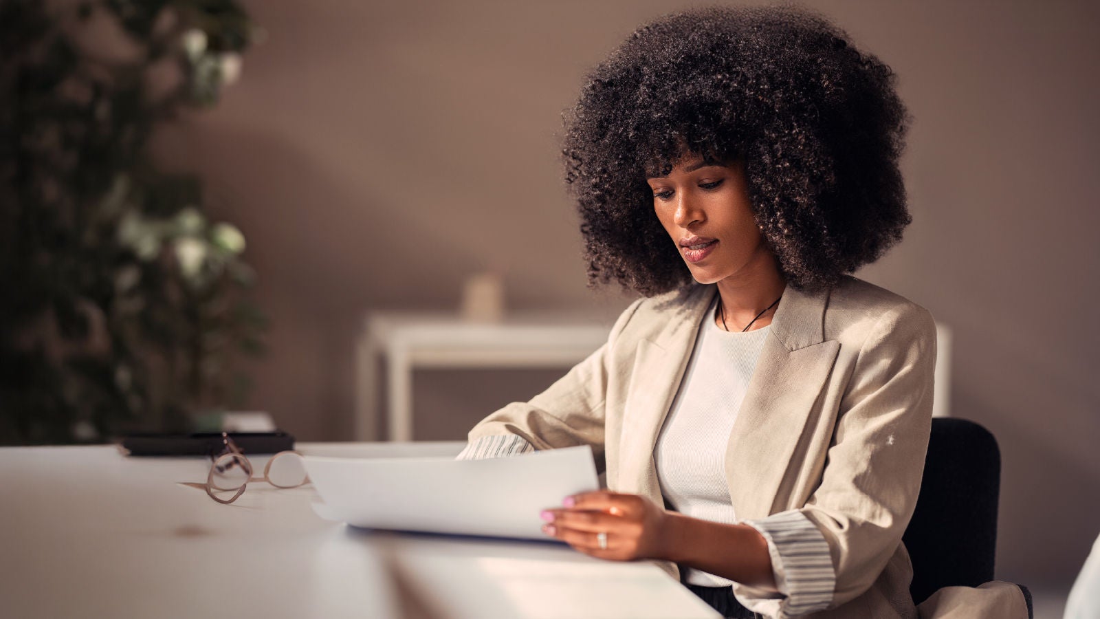 A woman in a beige blazer sits at a desk reviewing documents in a softly lit office, with reading glasses on the table and a blurred plant in the background.