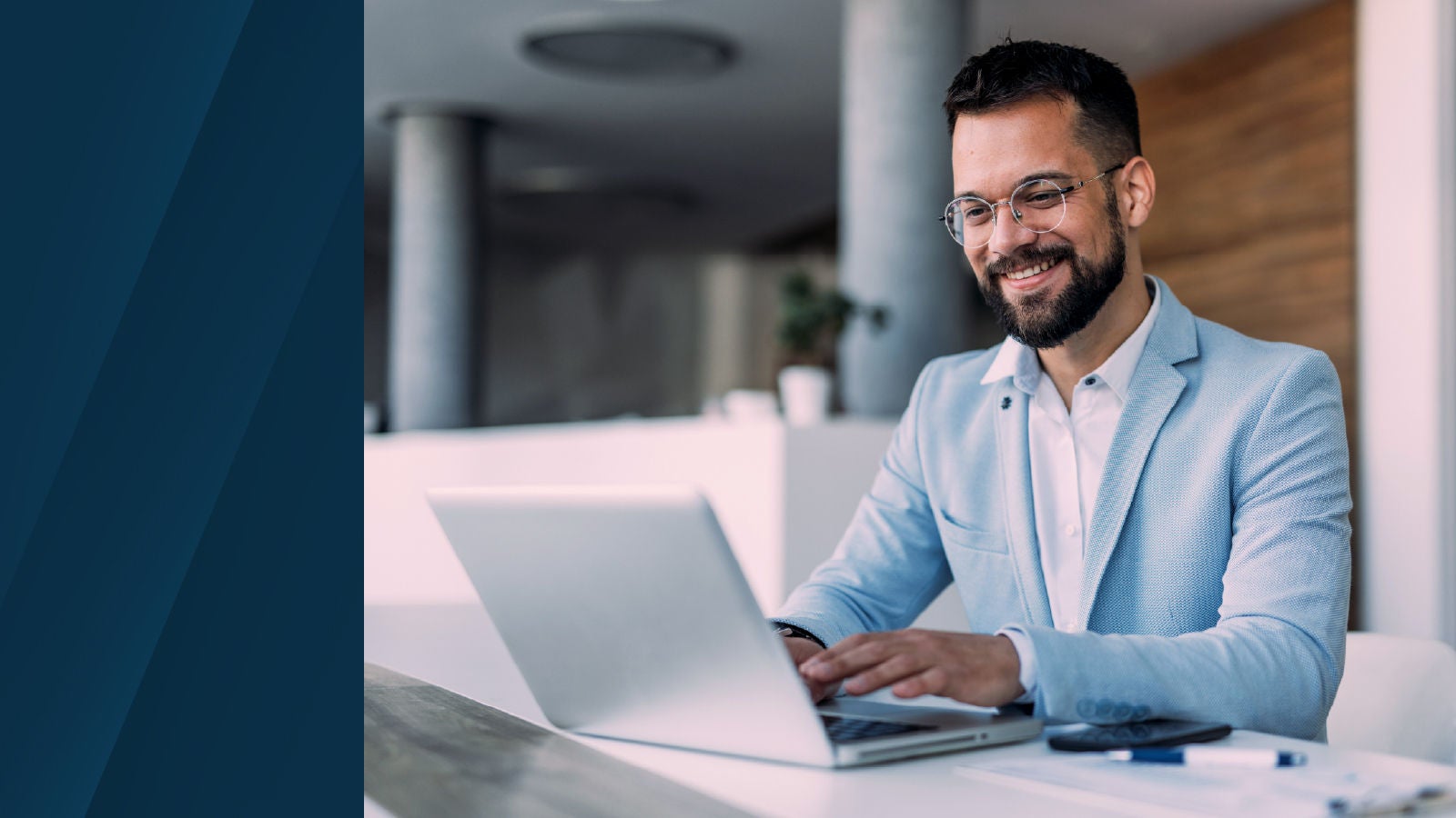 A smiling man in a light blue blazer sits at a white desk working on a laptop, with a paper and pen nearby in a modern office setting.