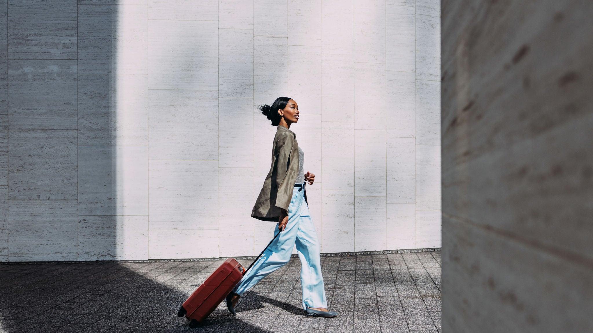 A woman walking outdoors while pulling a red suitcase along a light stone wall in daylight.