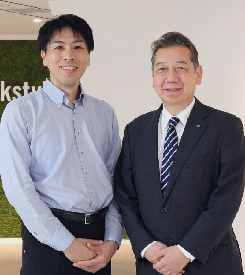 Two professional men in business attire standing together, smiling in a corporate office setting.