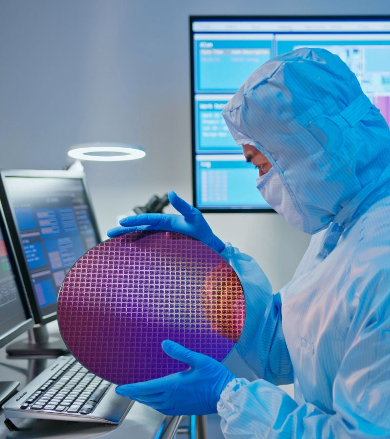 A technician in a cleanroom suit examining a semiconductor wafer in a high-tech lab with computer monitors displaying data.