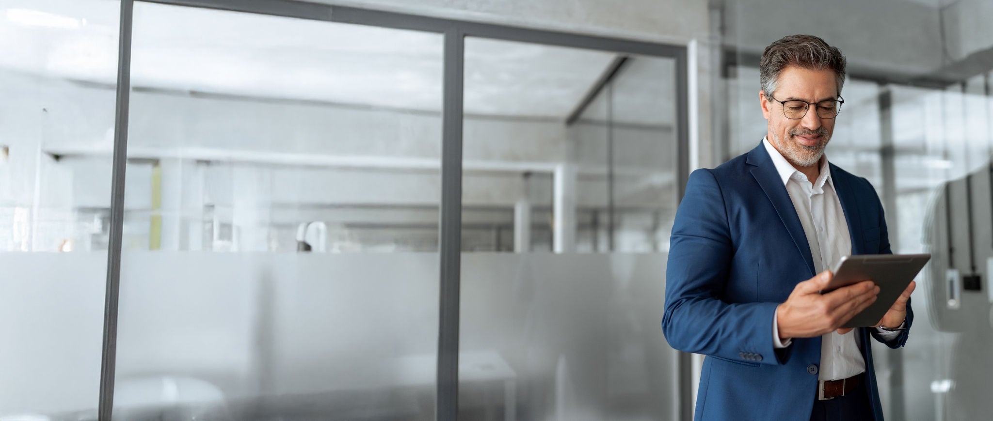 A professional in a blue suit stands in a modern office, smiling while using a tablet near glass walls.