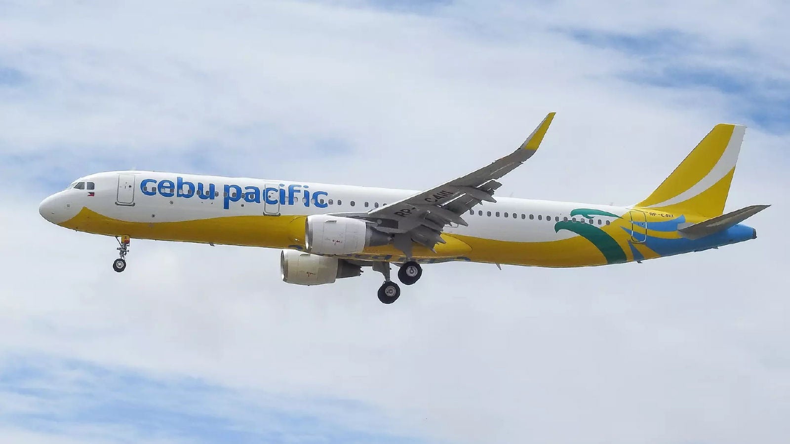 A Cebu Pacific passenger airplane in flight with landing gear extended, showing the white and yellow aircraft with the “cebu pacific” logo against a cloudy sky.
