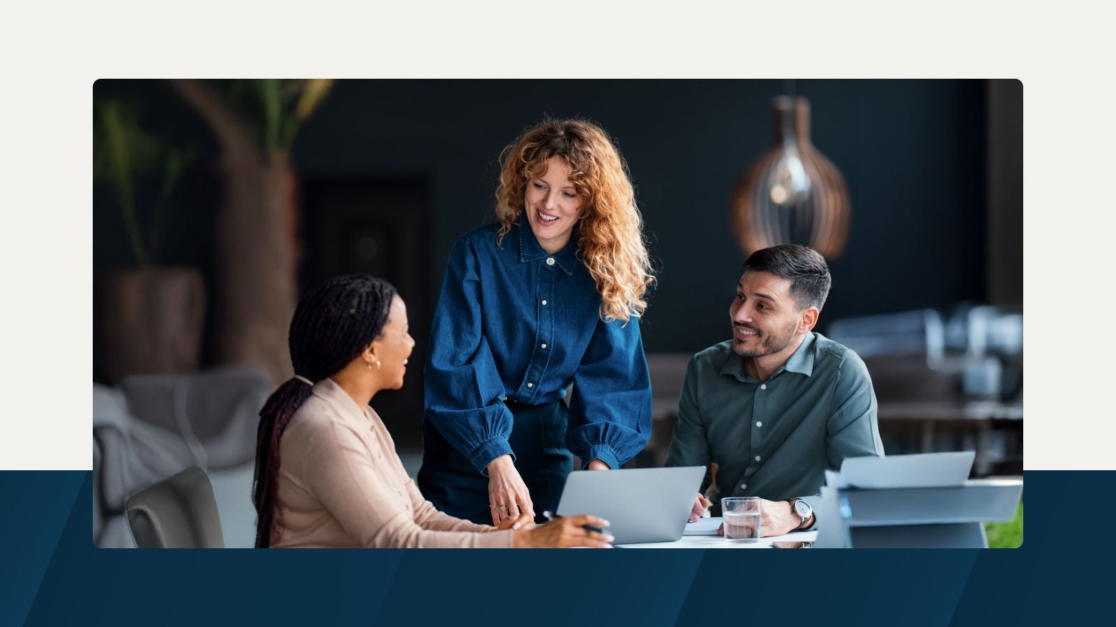 Three colleagues in a modern workspace talking and working together around a laptop, with a warm, softly lit background.