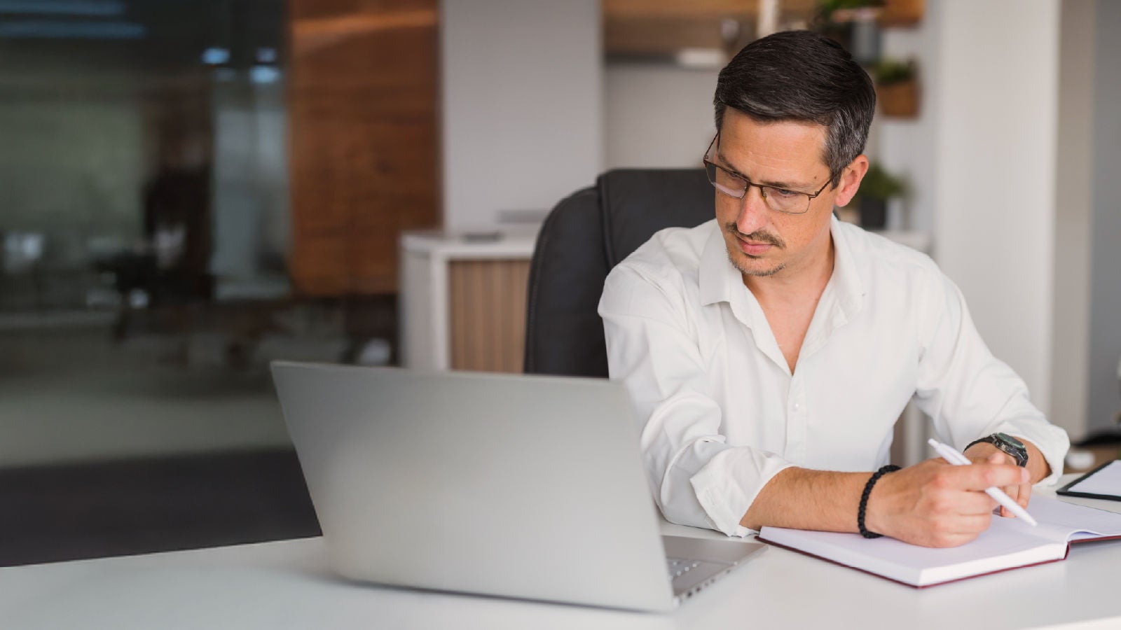 A professional in a white shirt works at a desk, taking notes while looking at a laptop in a contemporary office setting.