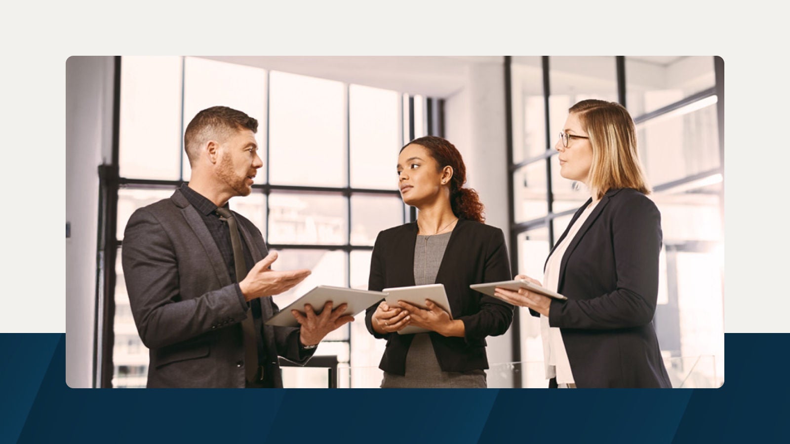 Three business professionals in formal attire discuss operating expense planning while holding tablets in a modern office with large windows.