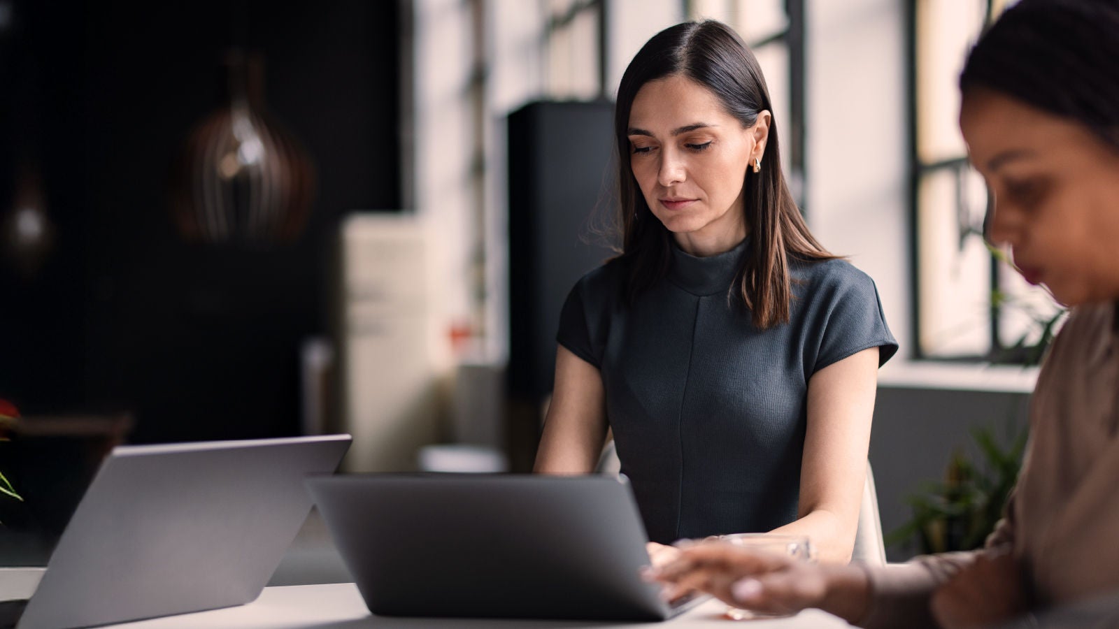 A focused woman in a dark dress works on a laptop at a modern office table, with another colleague blurred in the foreground and natural light streaming through large windows.