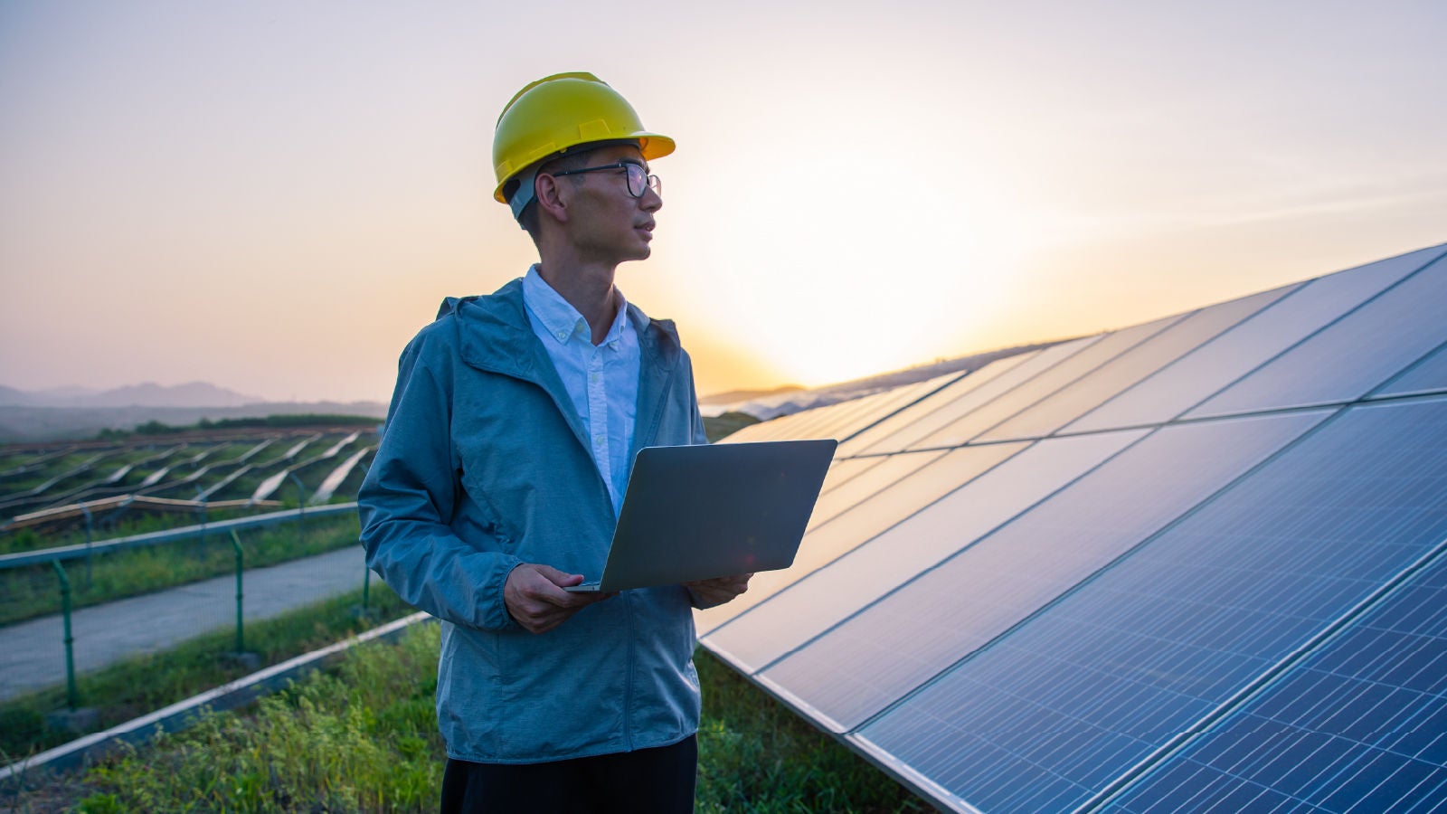 An engineer wearing a hard hat using a laptop while standing beside solar panels at sunset.
