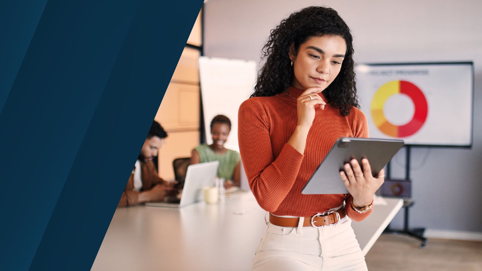 A woman standing in a modern meeting room reviewing a tablet, with colleagues seated at a table behind her and a presentation screen visible in the background.