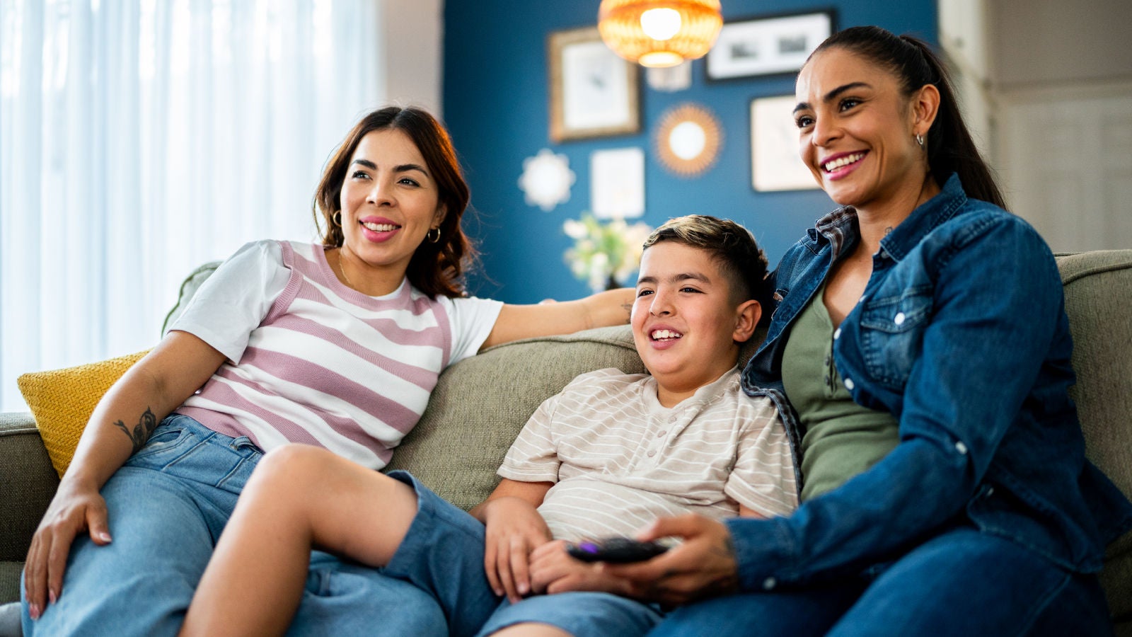 Two women and a young boy sit together on a sofa, smiling while watching TV. The boy holds a remote control. The room is warmly lit with framed artwork and decor on the wall in the background.
