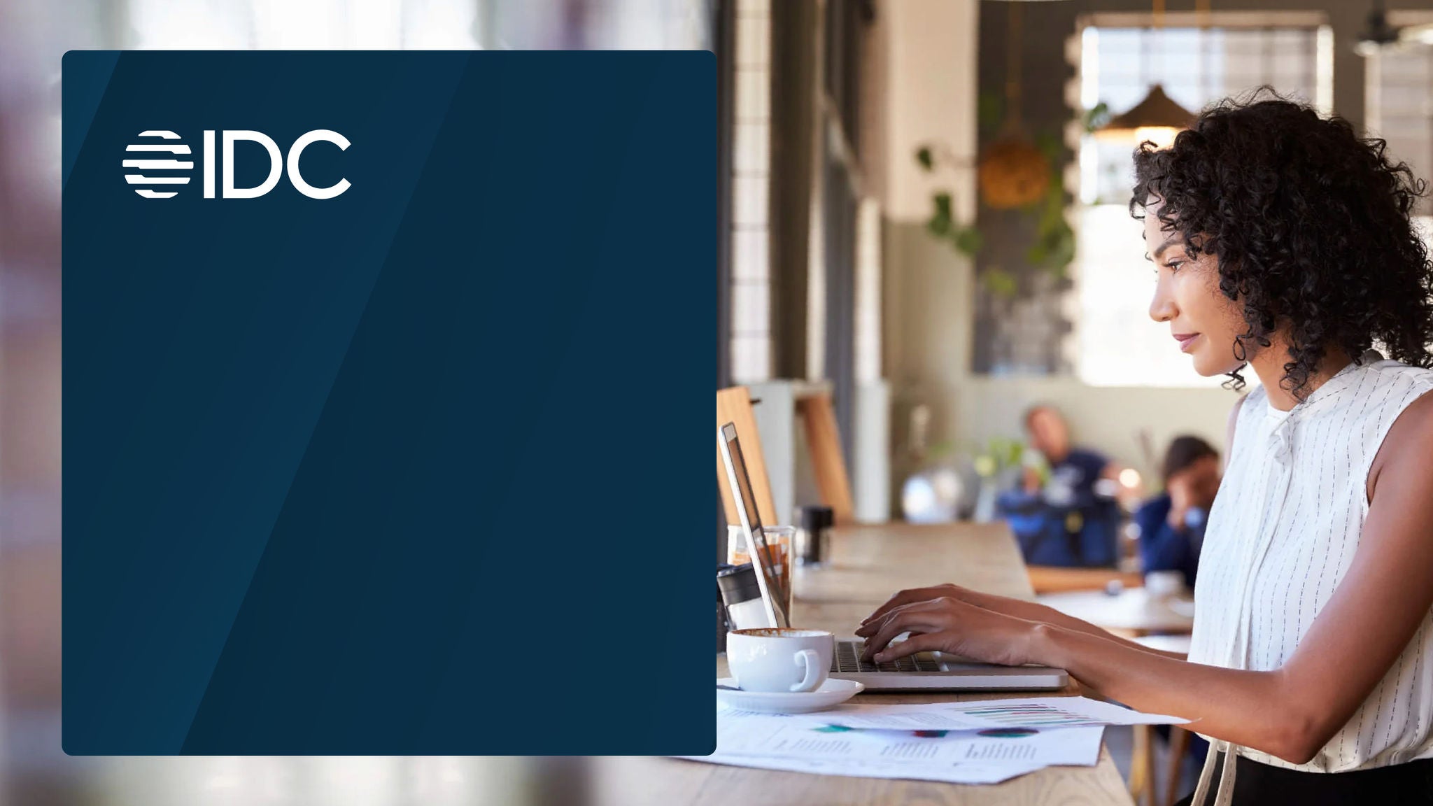 A woman working on a laptop at a café table, with an IDC logo overlay on the left side.