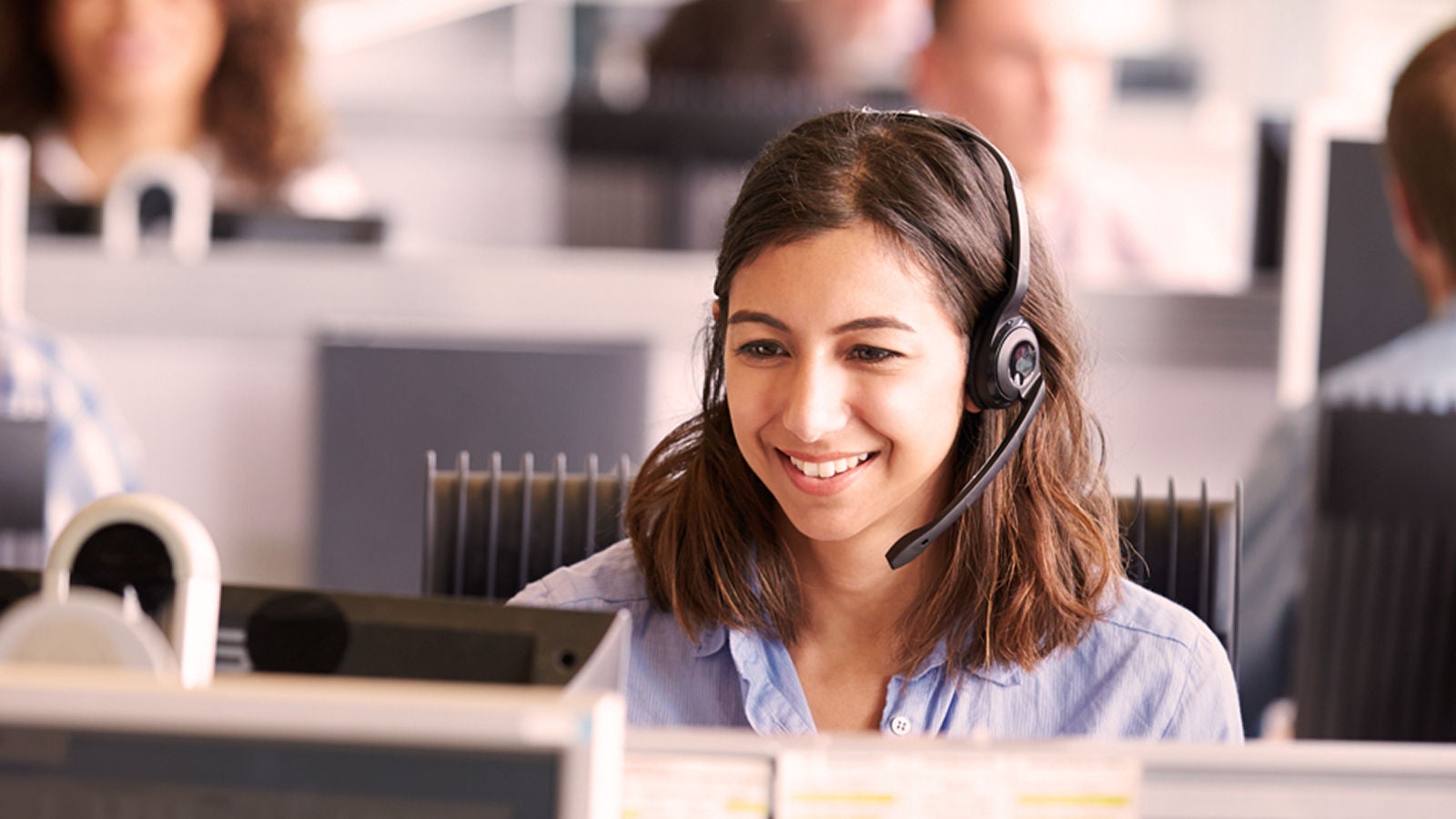 A woman smiling while wearing a headset, seated in a modern contact center environment.