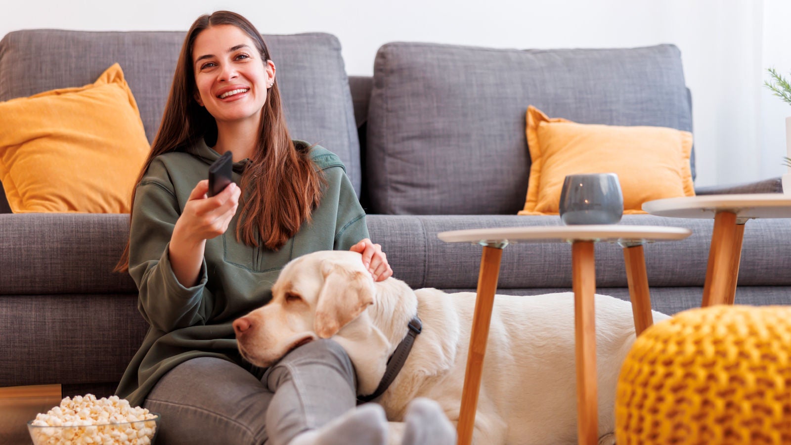A woman sits on a sofa with a dog on her lap, holding a remote and smiling while watching TV. A bowl of popcorn rests on the floor beside her. A coffee table and a gray sofa with yellow pillows are in the background.