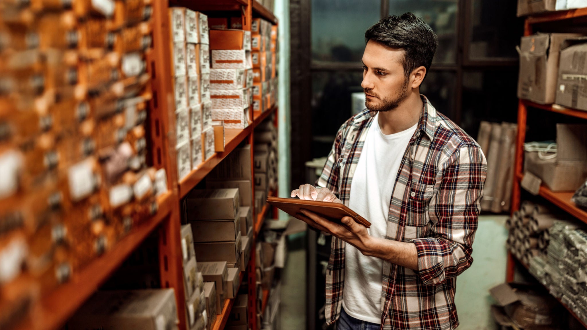 A warehouse worker checking inventory on a tablet while standing between shelves stocked with boxed goods.