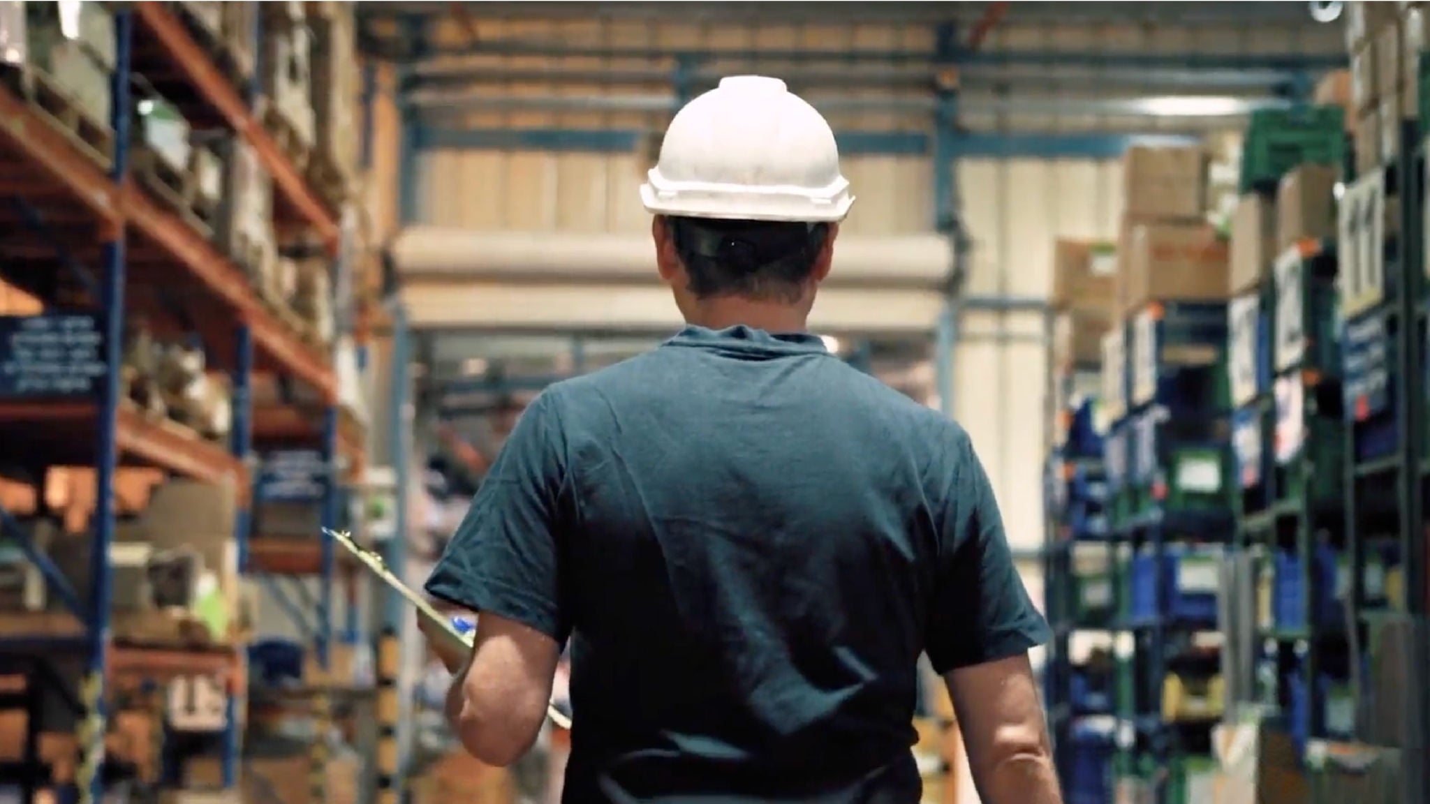 A worker in a warehouse wearing a white hard hat, holding a clipboard, and walking down an aisle lined with shelves of boxes and containers.