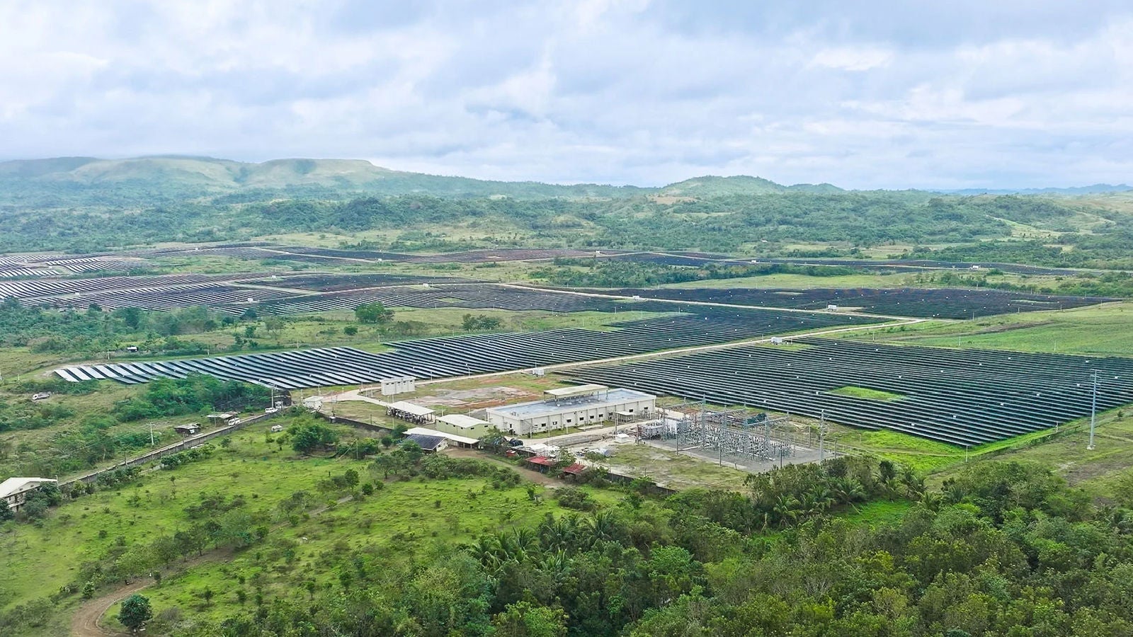 Aerial view of a large solar farm with rows of solar panels spread across a green landscape with hills in the background.