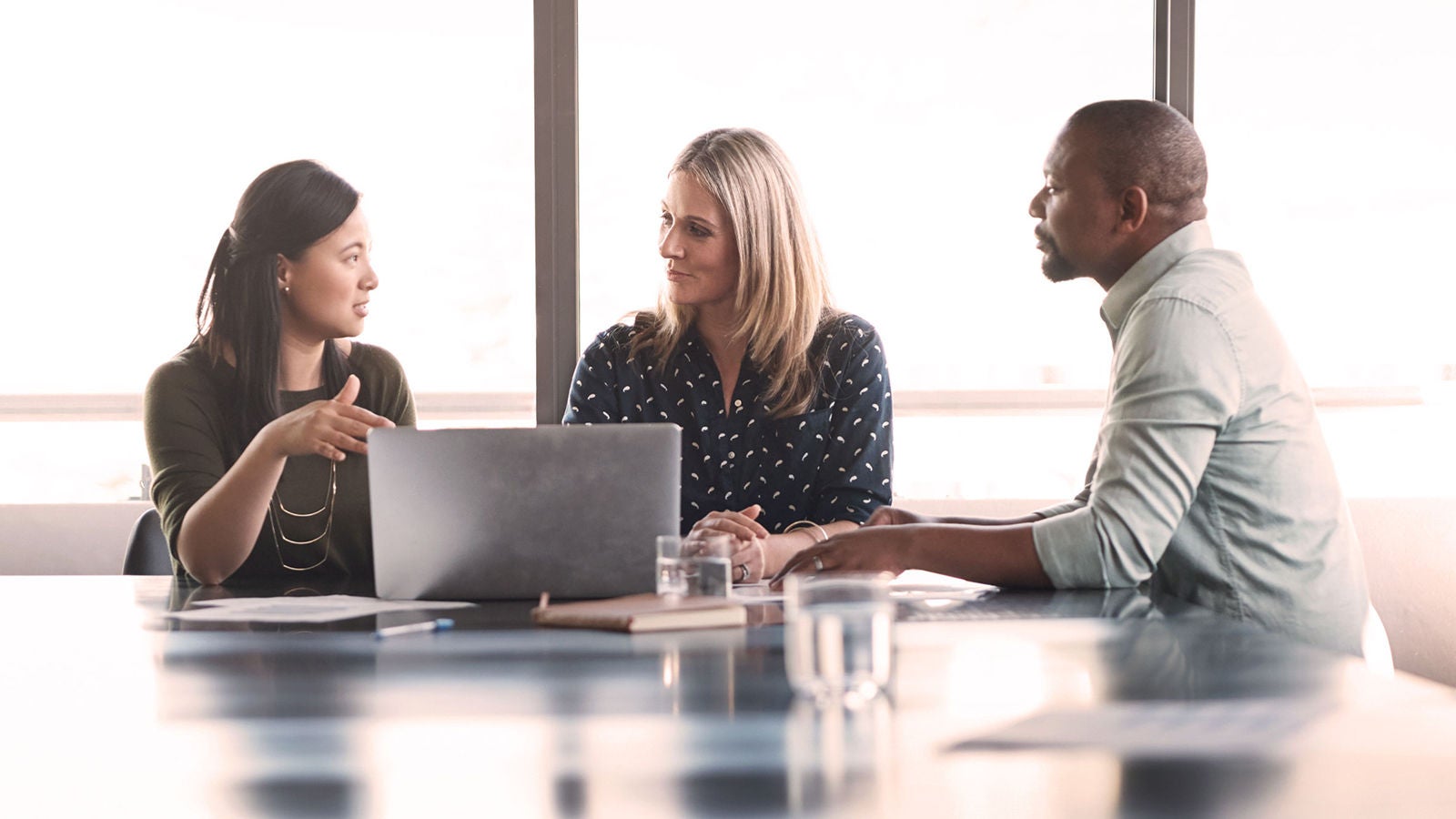 Three professionals engaged in a discussion at a conference table, with a laptop open and documents spread out, in a bright office setting.
