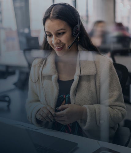 A woman wearing a headset smiles while working on a laptop in a modern office environment.