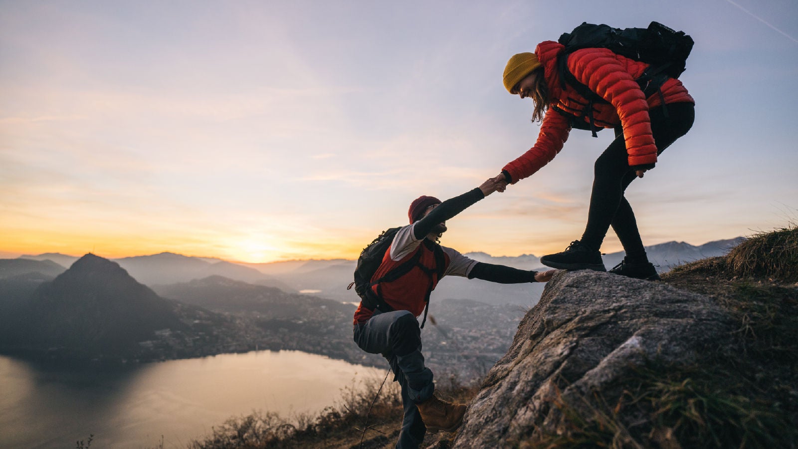 Two hikers on a mountain at sunset, one helping the other climb up a rocky ledge overlooking a lake and valley.