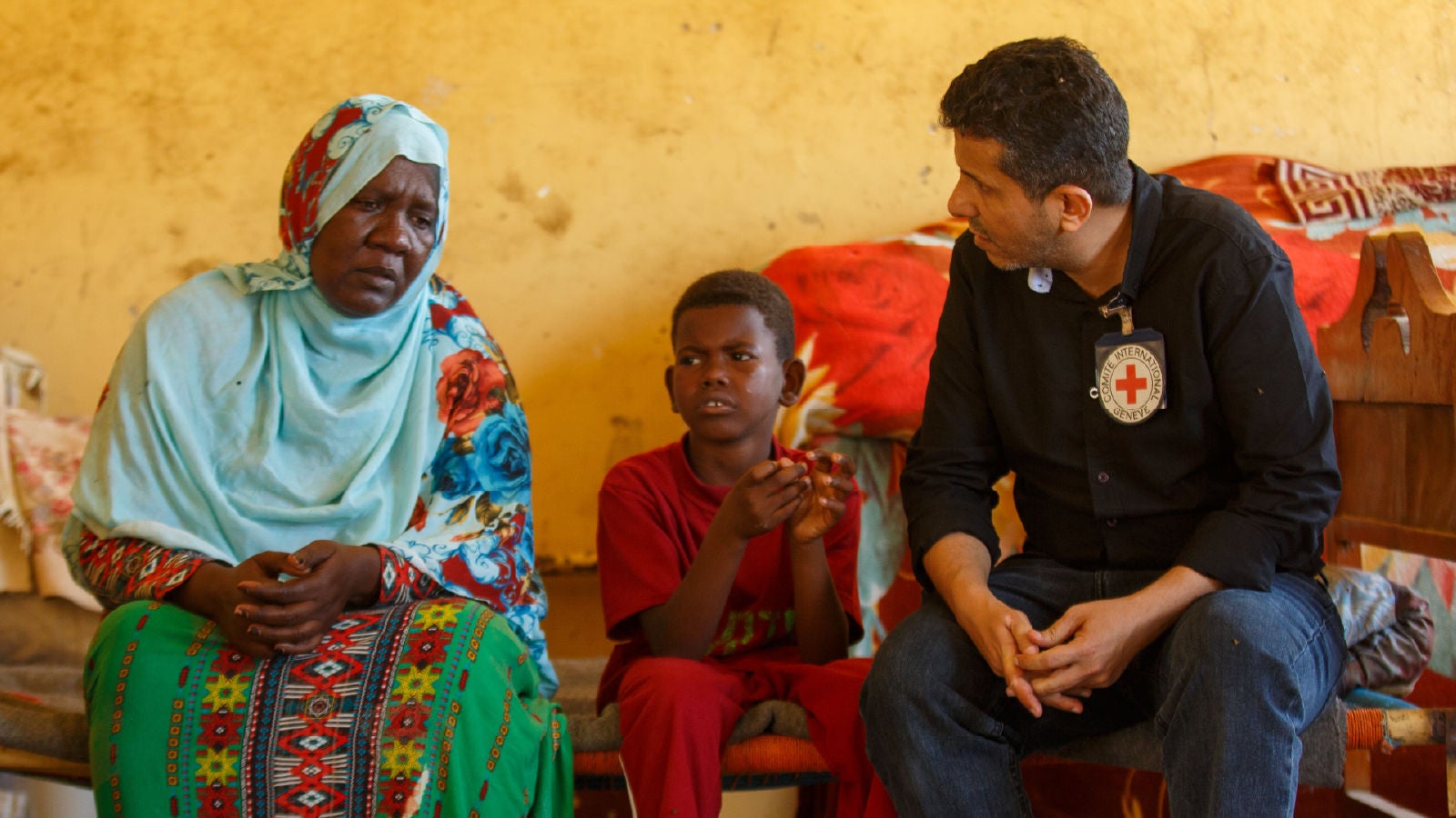 An ICRC staff member speaks with a woman in a blue headscarf and a boy in red clothing inside a modest home.