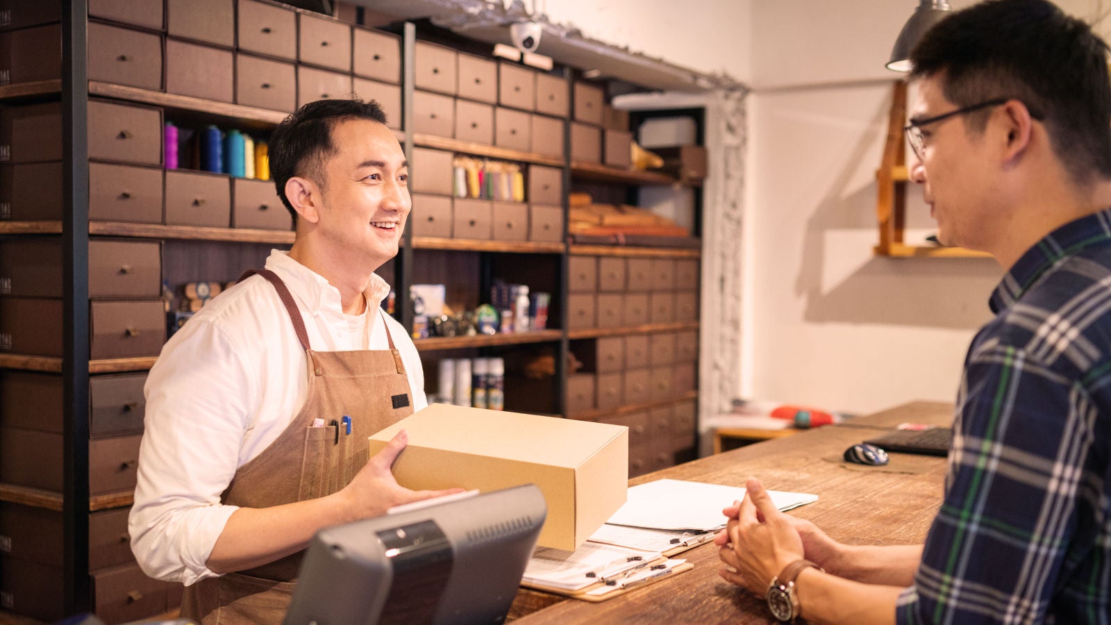 Shop employee holding a parcel while speaking with a customer across the counter.
