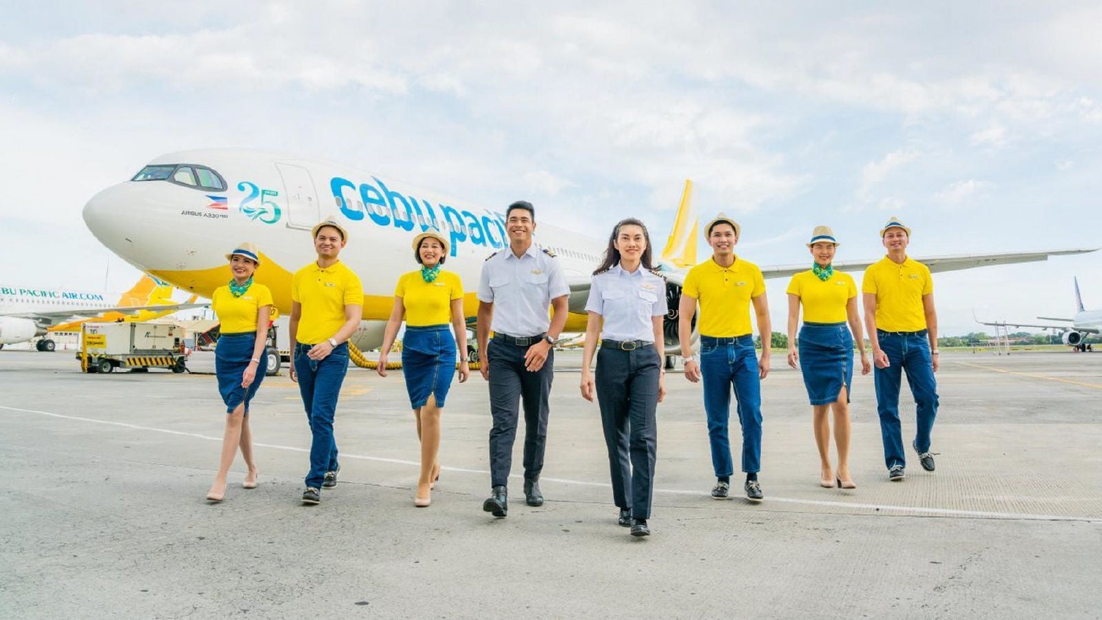 A group of Cebu Pacific airline crew members walking on an airport tarmac in front of a parked aircraft, wearing yellow uniforms, with the airplane visible in the background.