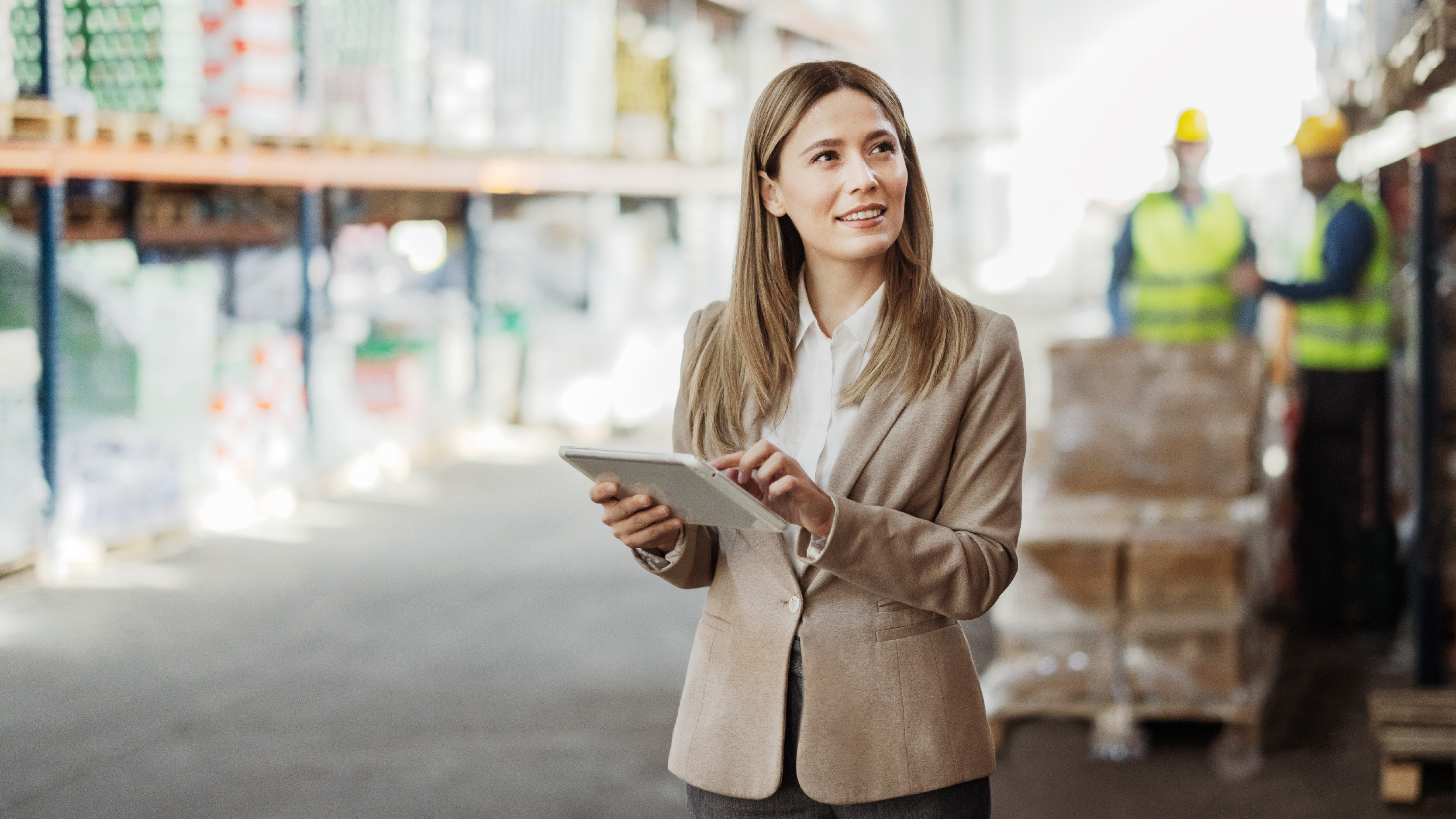 A business professional using a tablet inside a warehouse, with inventory shelves and workers wearing safety vests visible in the background.