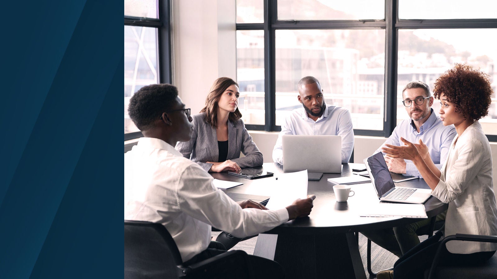 A diverse group of professionals in discussion around a conference table, with laptops and papers, in a modern office with large windows.