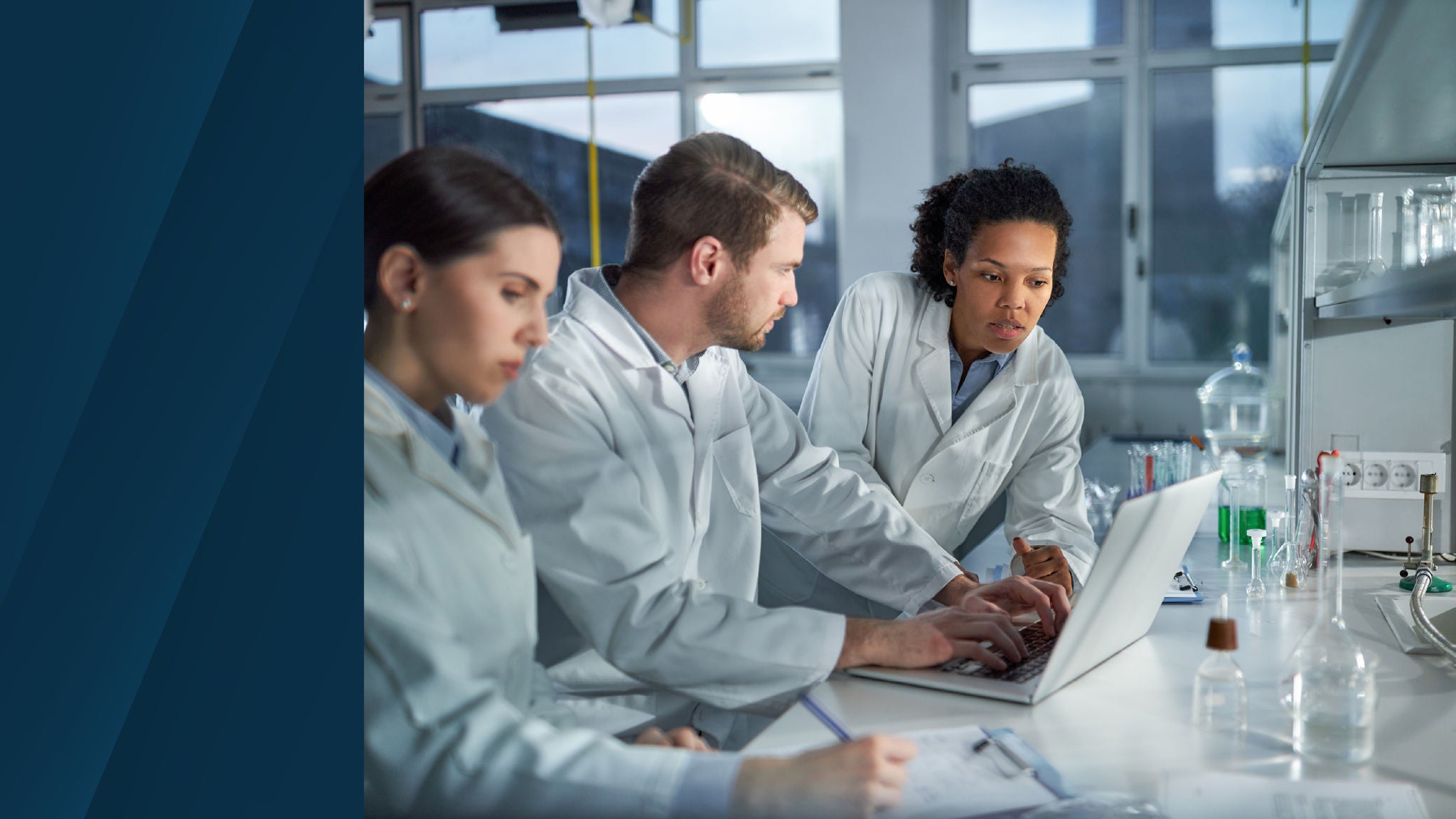 Three lab professionals in white coats collaborate in a modern laboratory setting, focused on a laptop with scientific glassware visible on the counter.