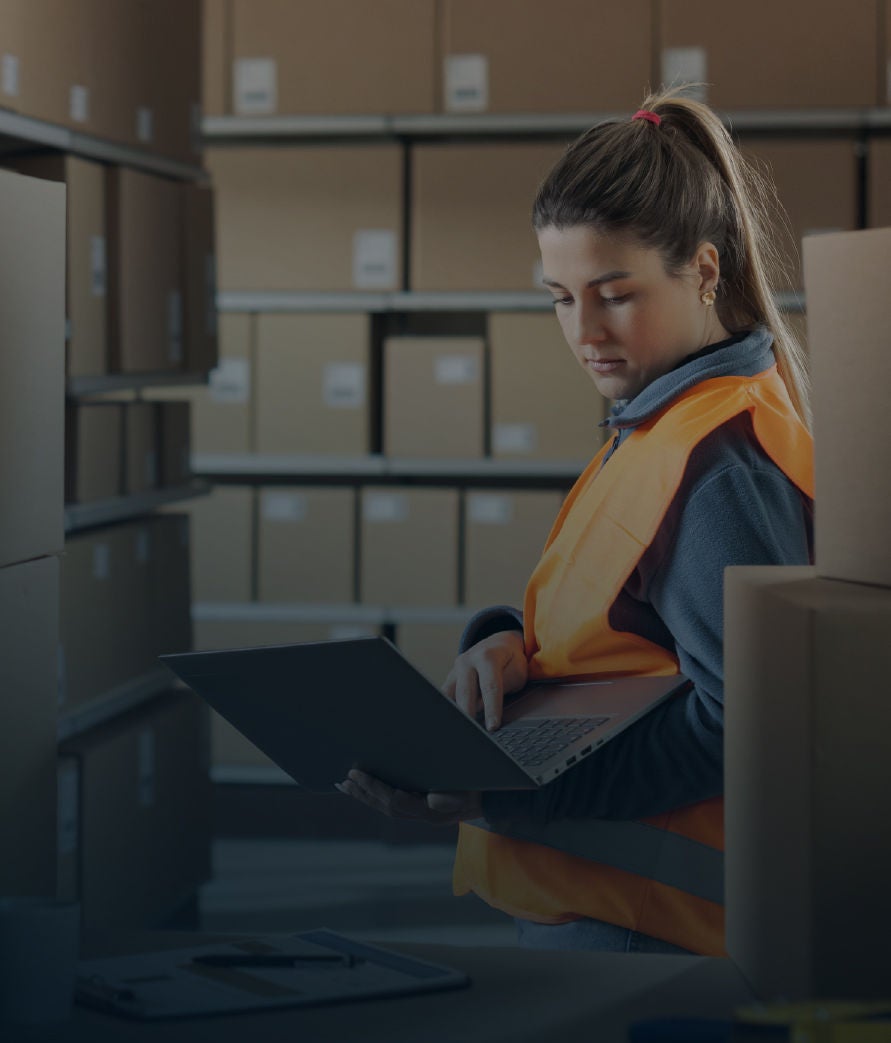 A warehouse worker wearing a high-visibility vest reviews inventory on a laptop while standing among stacked cardboard boxes in a storage facility.