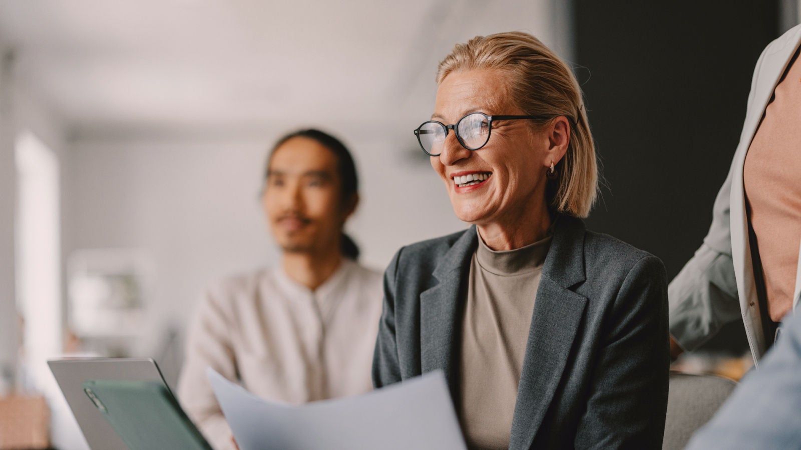 A smiling businesswoman wearing glasses and a gray blazer holds a document while engaged in a discussion. A blurred colleague is visible in the background.