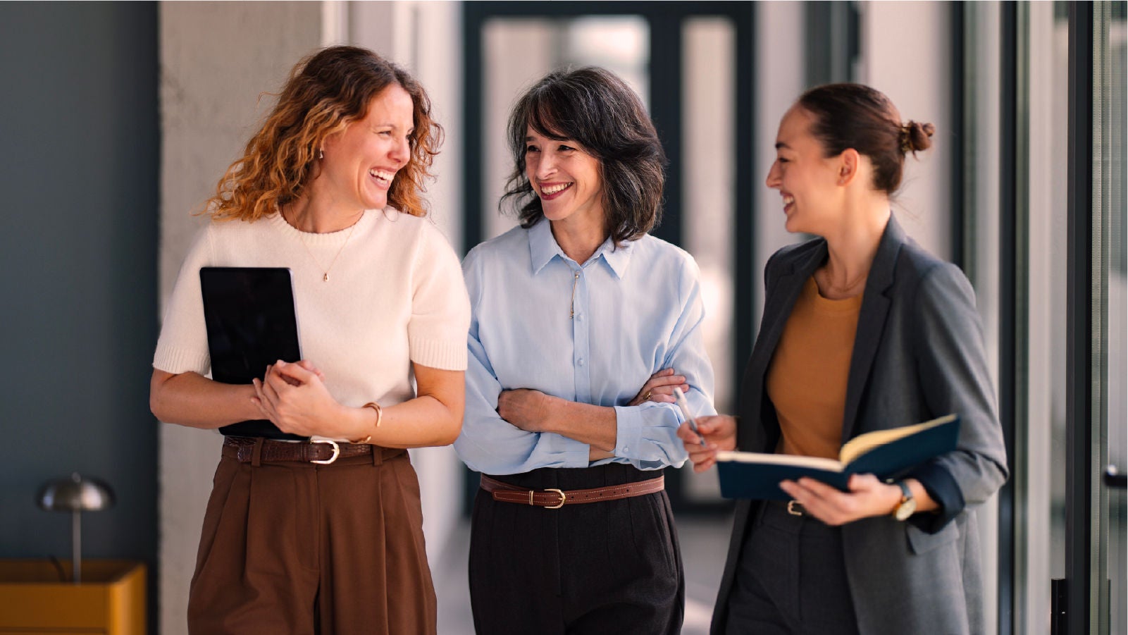 Three women in business attire walking together in an office hallway, smiling and holding a tablet and notebook.