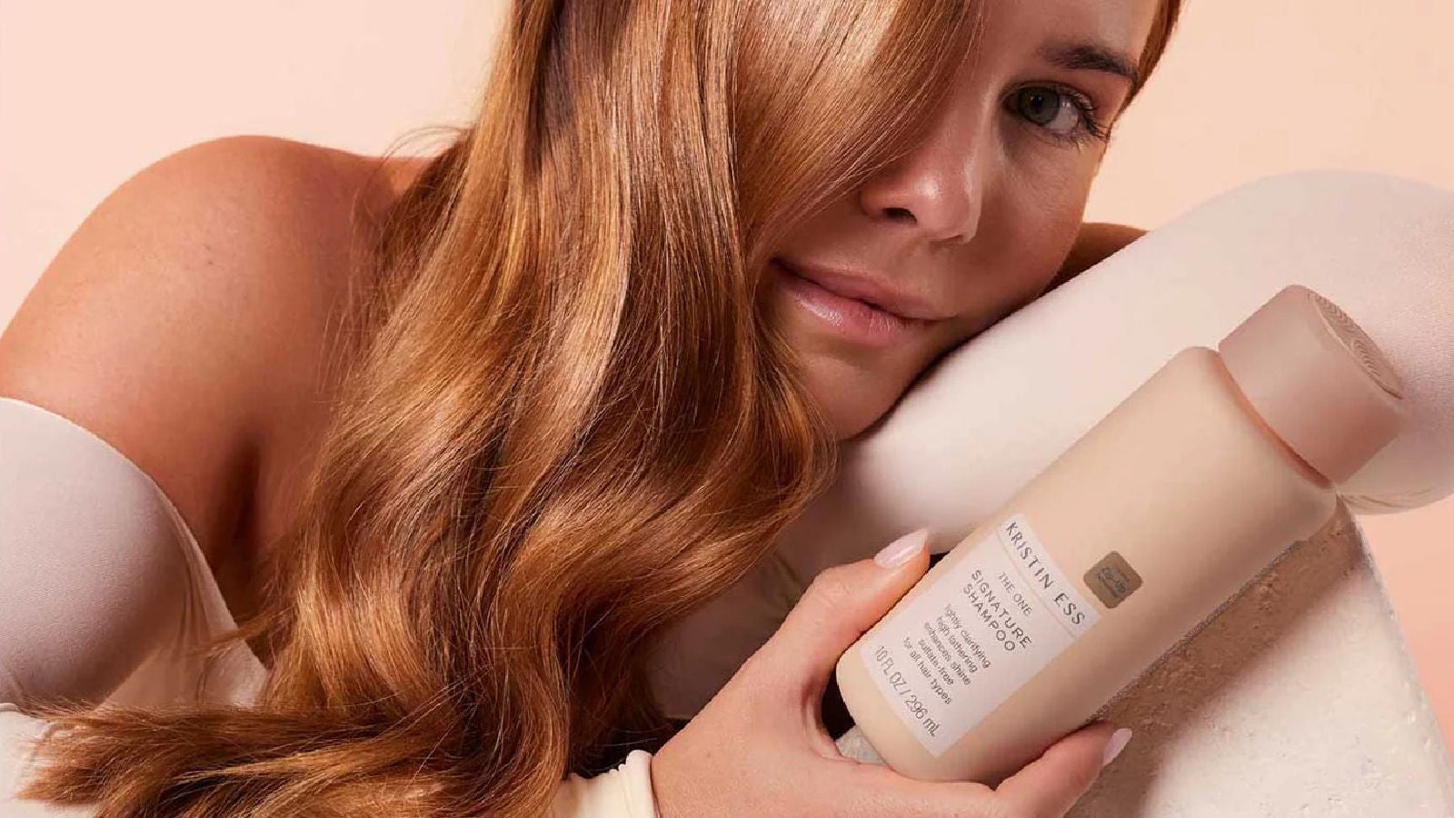 A close-up of a woman resting her head on a cushion while holding a shampoo bottle, with warm lighting highlighting her hair and the product packaging.