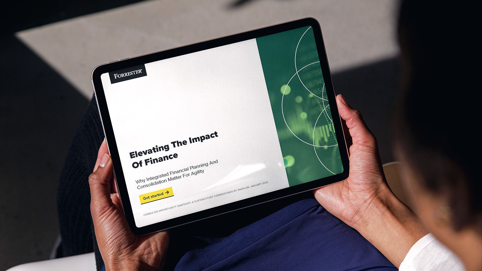 A person holding a tablet displaying a Forrester report titled “Elevating The Impact Of Finance: Why Integrated Financial Planning And Consolidation Matter For Agility,” with a “Get started” button and green graphic elements on the right side of the screen.
