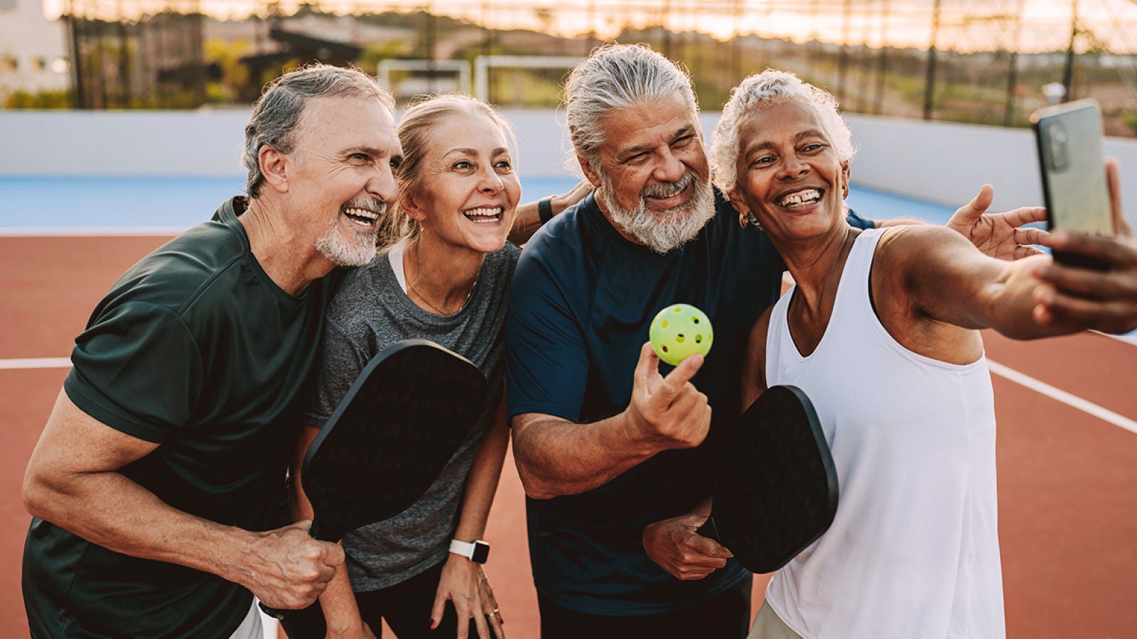 Four older adults taking a selfie while holding pickleball paddles and a ball on an outdoor court.