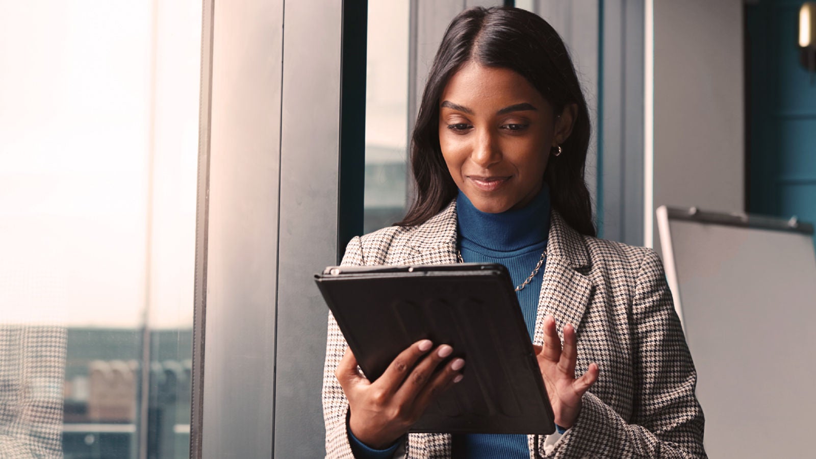 A woman in a checkered blazer and blue turtleneck smiles while using a tablet, standing near a window with light streaming in.