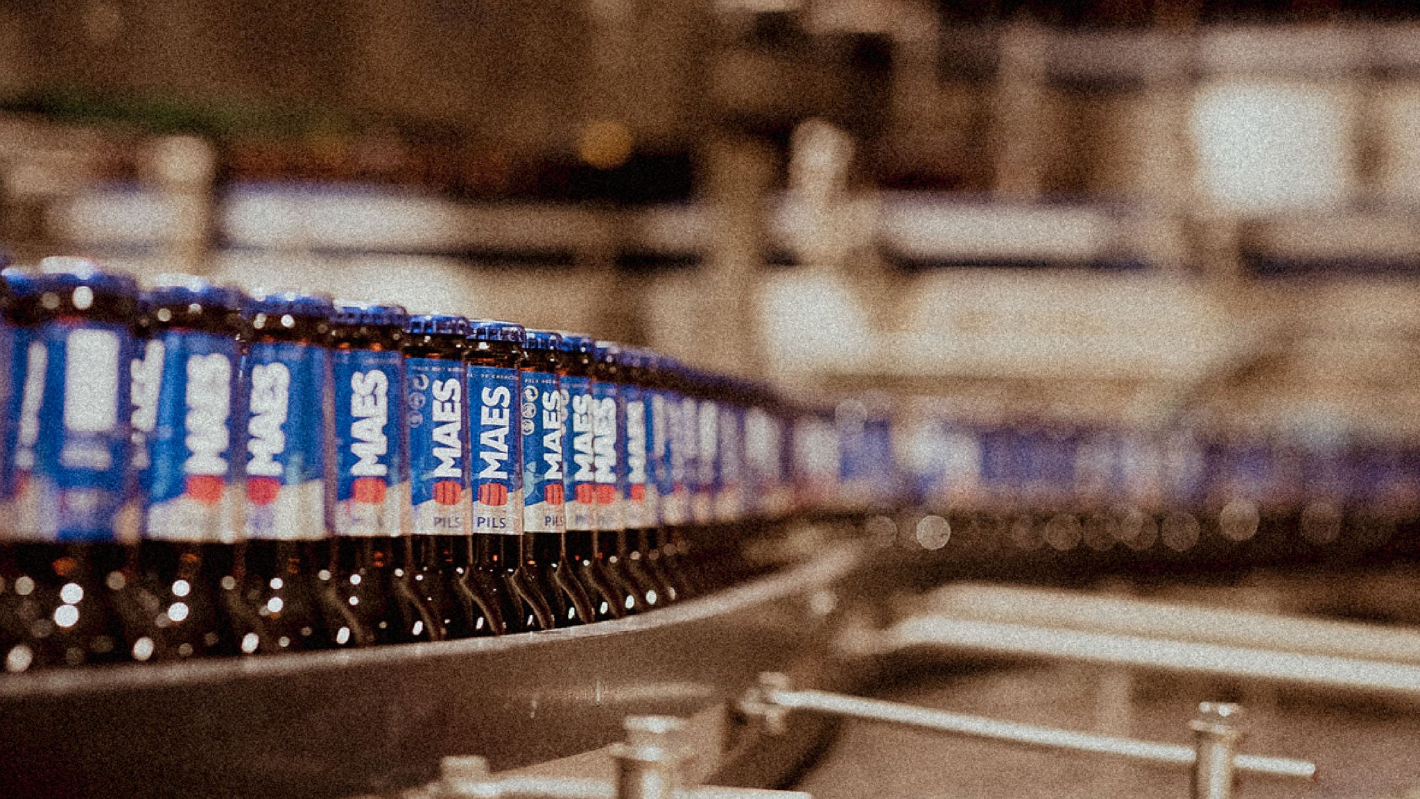 Rows of bottled beer moving along a production line inside a brewery, with machinery and conveyor belts in the background.