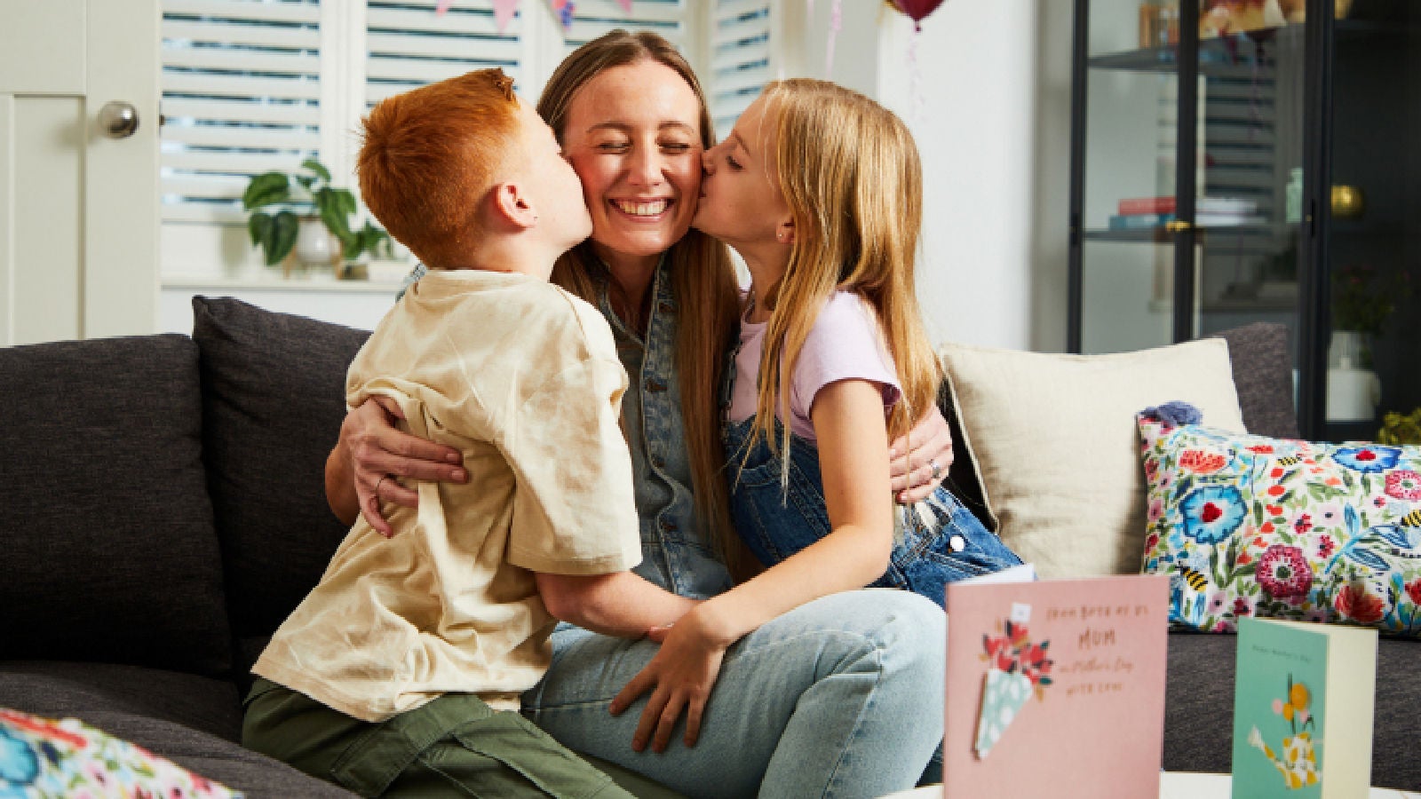 A smiling adult on a sofa being kissed on both cheeks by two children, with greeting cards and colorful pillows visible in the cozy room.