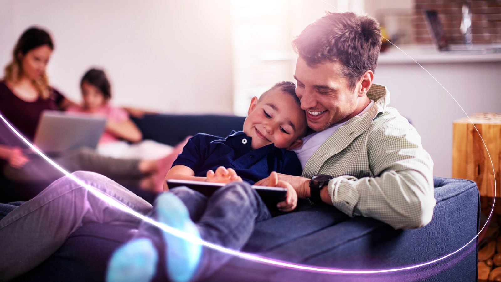 Father and young child sitting together on a couch and smiling while using a tablet, with a softly blurred family scene in the background.