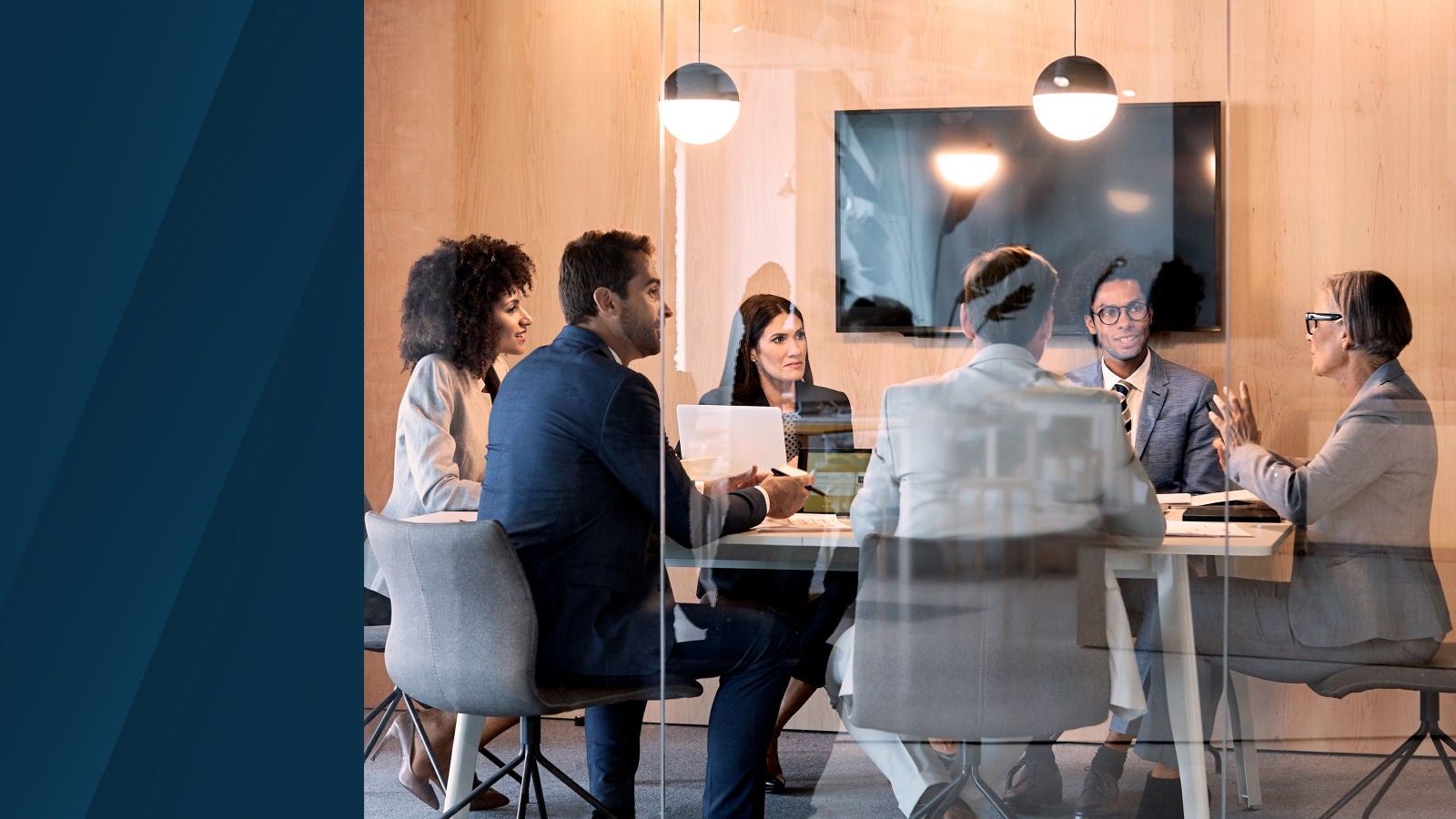 A group of six professionals engaged in a business meeting inside a glass-walled conference room, seated around a table with laptops and notes.